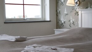 A neatly made bed with a folded towel and a remote control, a window with a cross pattern, and a wall-mounted lamp.