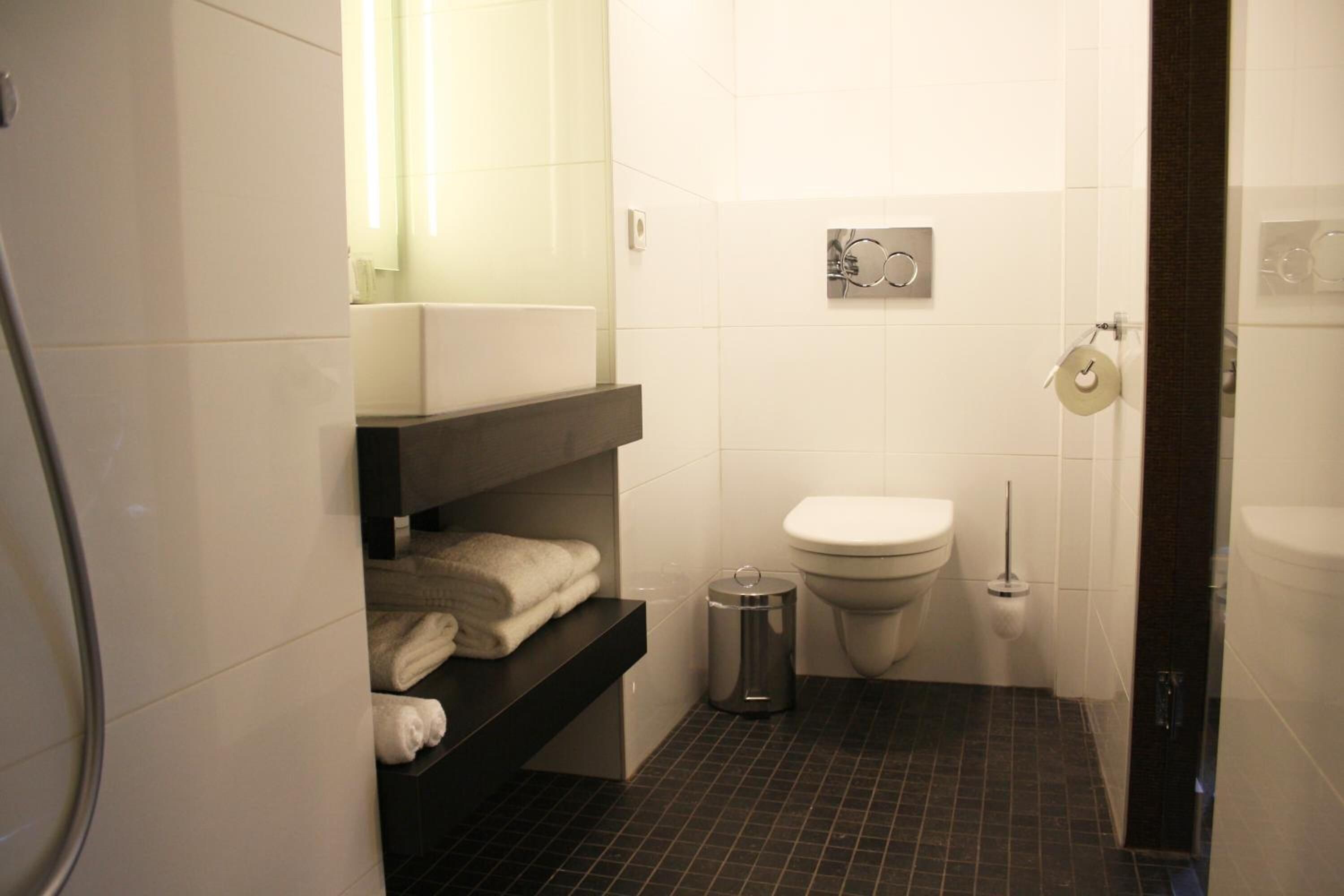 A small, modern bathroom with a toilet, a trash can, and a shelf with towels.