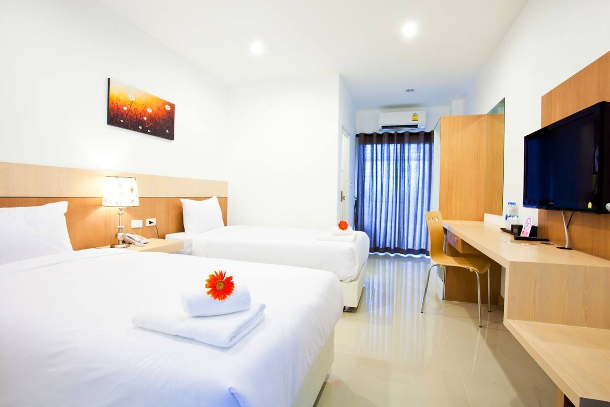A hotel room with two beds, a desk, a TV, and an air conditioning unit.
