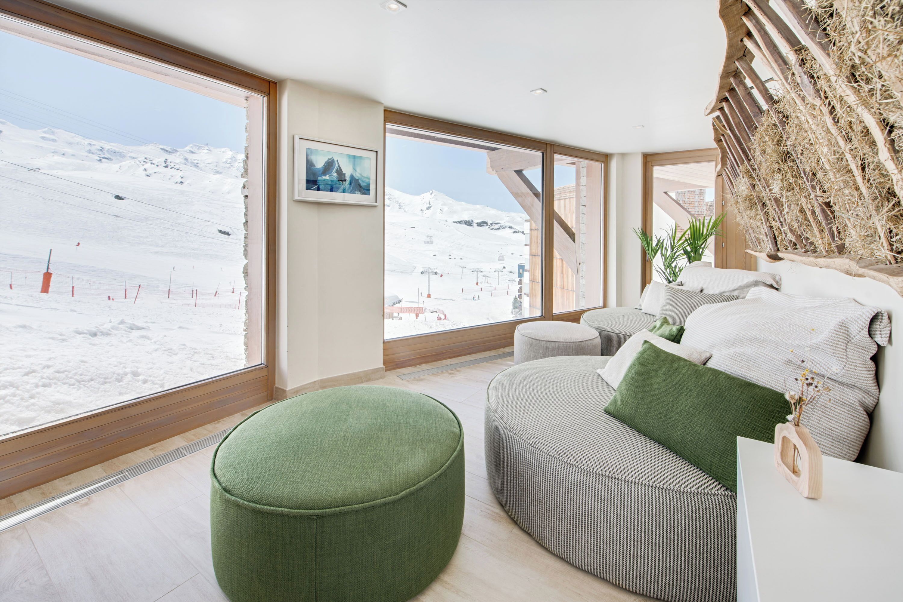 A modern room with a large window offering a snowy mountain view, a green ottoman, a striped sofa, and a wooden wall decor.