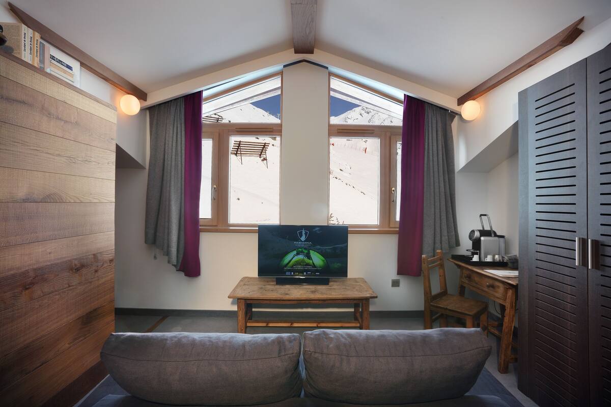 Ein gemütliches Zimmer mit Fernseher auf einem Holztisch, einer Couch und einem Fenster mit Vorhängen.