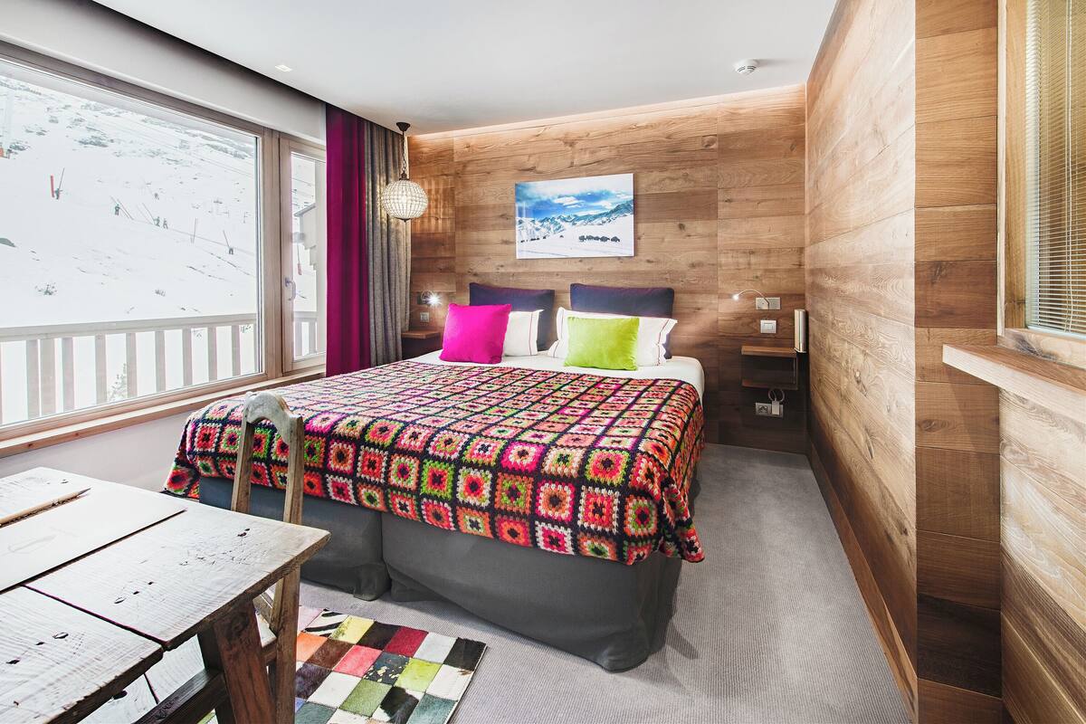 Ein gemütliches Zimmer mit einem großen Bett, einer Holzwand, einem Fenster mit Blick auf eine verschneite Landschaft und einem Tisch mit einer farbenfrohen Stegdecke.