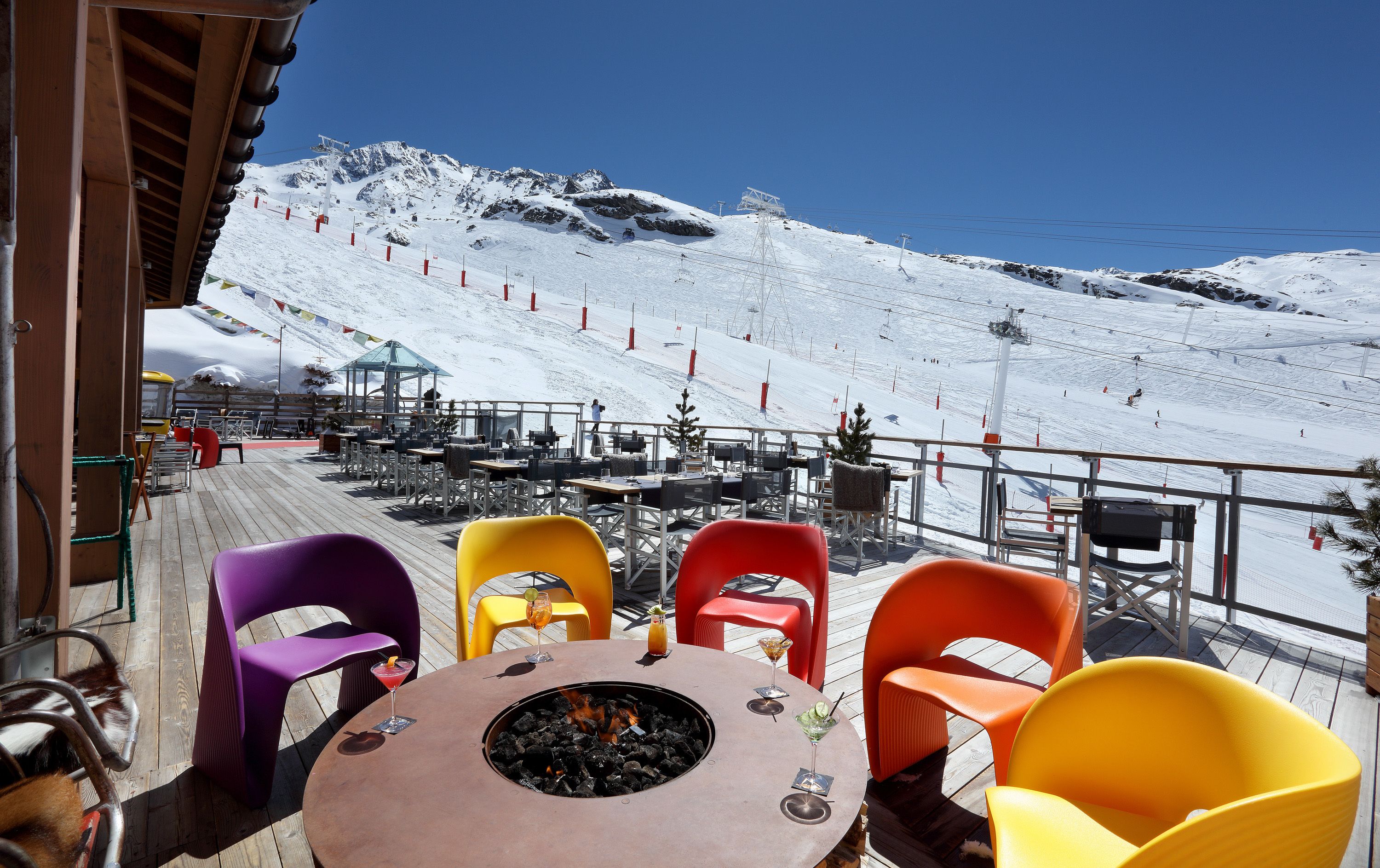 A wooden deck with colorful chairs and a round table with a fire pit, overlooking a snowy mountain slope with ski lifts.