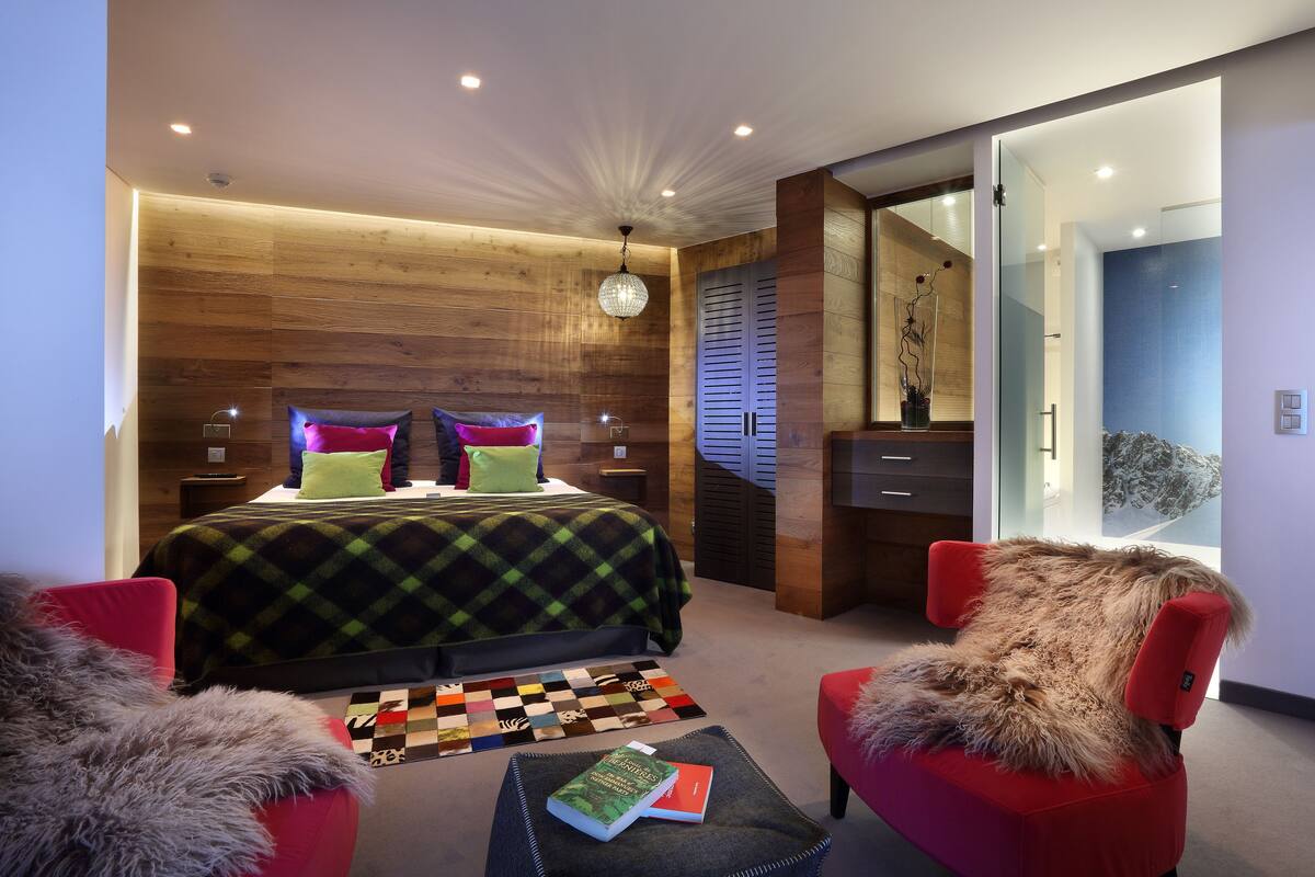 Ein modernes Schlafzimmer mit einer Holzwand, einem Bett mit bunten Kissen, einem roten Sessel und einem durchsichtigen Badezimmer.
