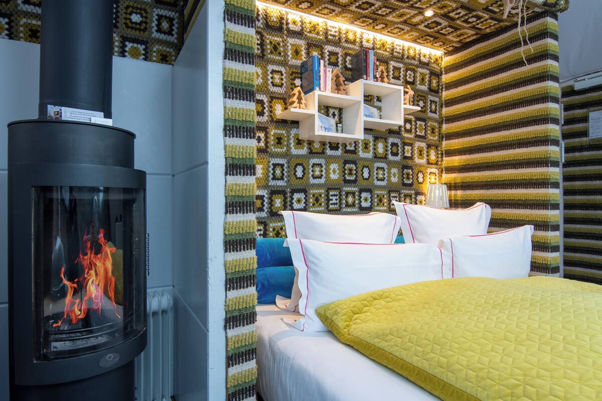 Ein gemütliches Zimmer mit Kamin, einem Bett mit weißer und gelber Bettwäsche und einer Wand mit einem geometrischen Muster.