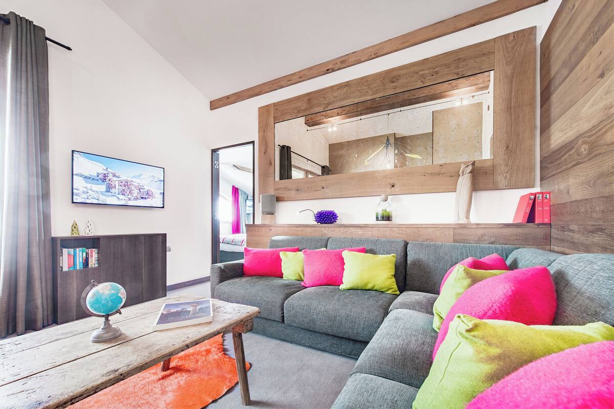 Ein modernes Wohnzimmer mit einem grauen Ecksofa, bunten Kissen, einer Holzzieldecke und einem Globus auf einem Holztisch.