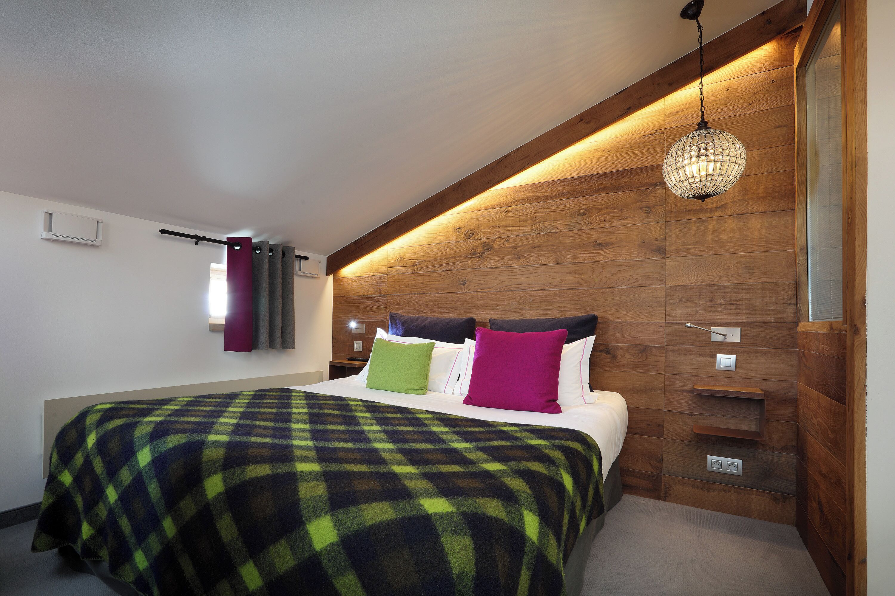 Une chambre à coucher avec une tête de lit en bois, un grand lit recouvert d’une couverture à carreaux et un lustre en cristal suspendu au plafond.