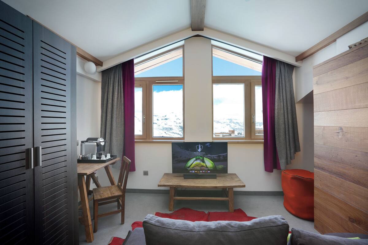 Ein gemütliches Zimmer mit schräg stehender Decke, einem Holztisch mit Kaffeemaschine, einem Fernseher und einem Fenster mit Vorhängen.