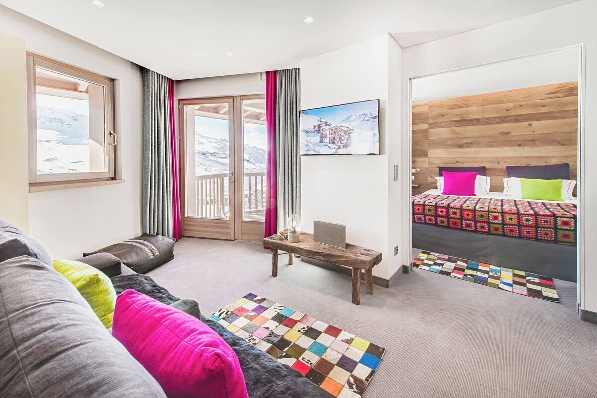 Ein modernes Hotelzimmer mit großem Fenster, einer gemütlichen Sitzecke, einem Bett mit Holz Kopfteil und einem farbenfrohen Teppich.
