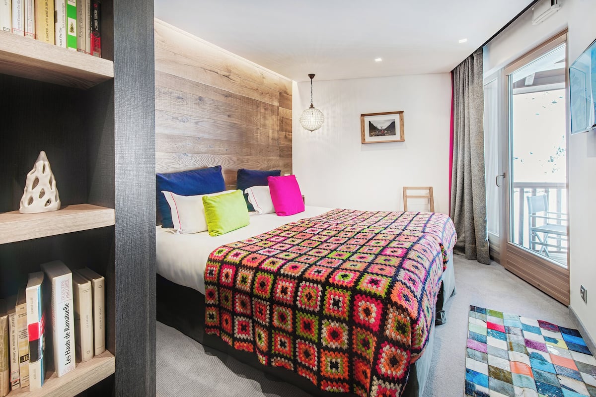Ein modernes Schlafzimmer mit einem großen Bett, einem Bücherregal, einem gerahmten Bild an der Wand und Blick durch ein Fenster.
