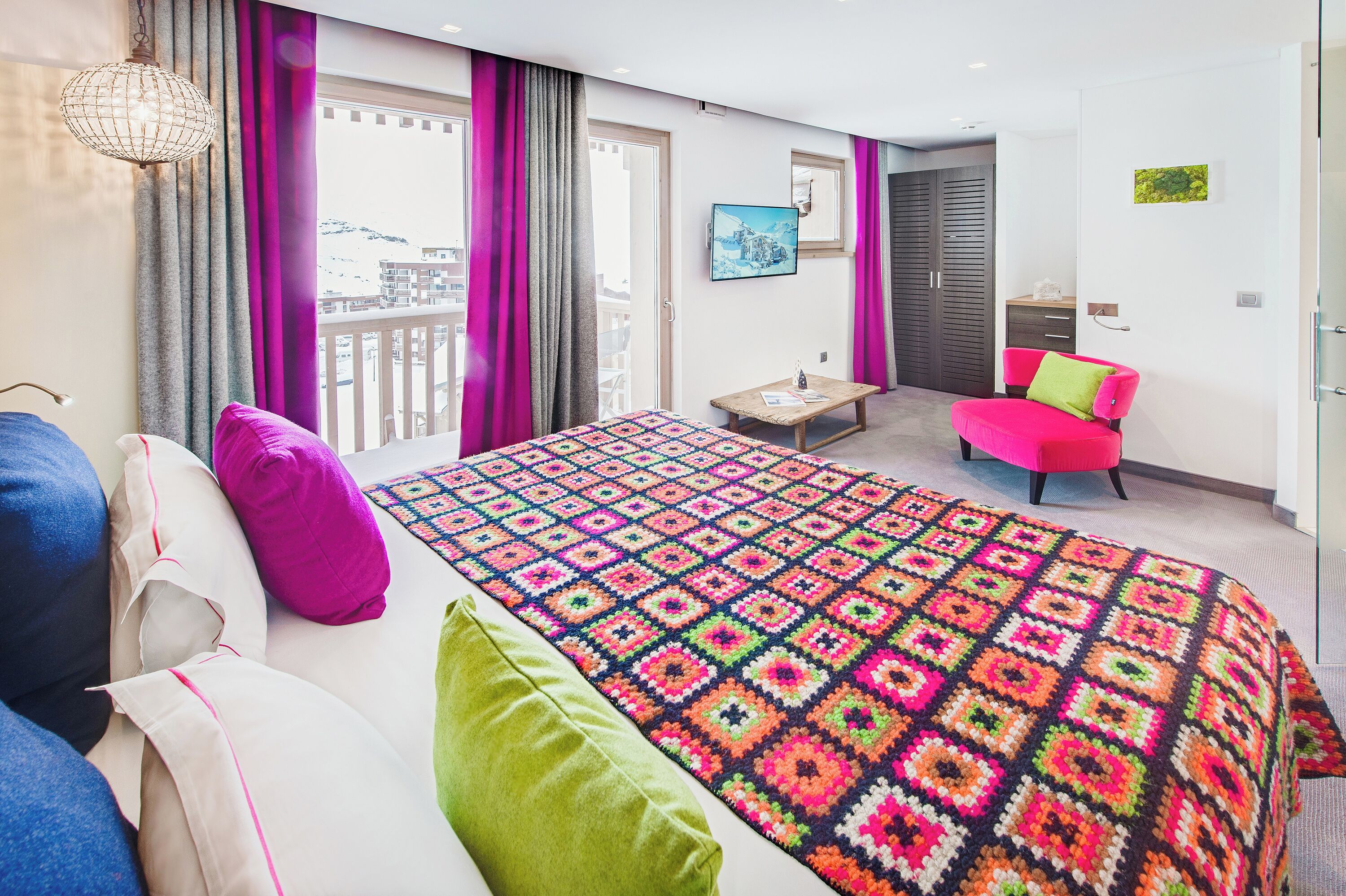 A bedroom with a colorful blanket, a bed, a chair, and a view of the outside.