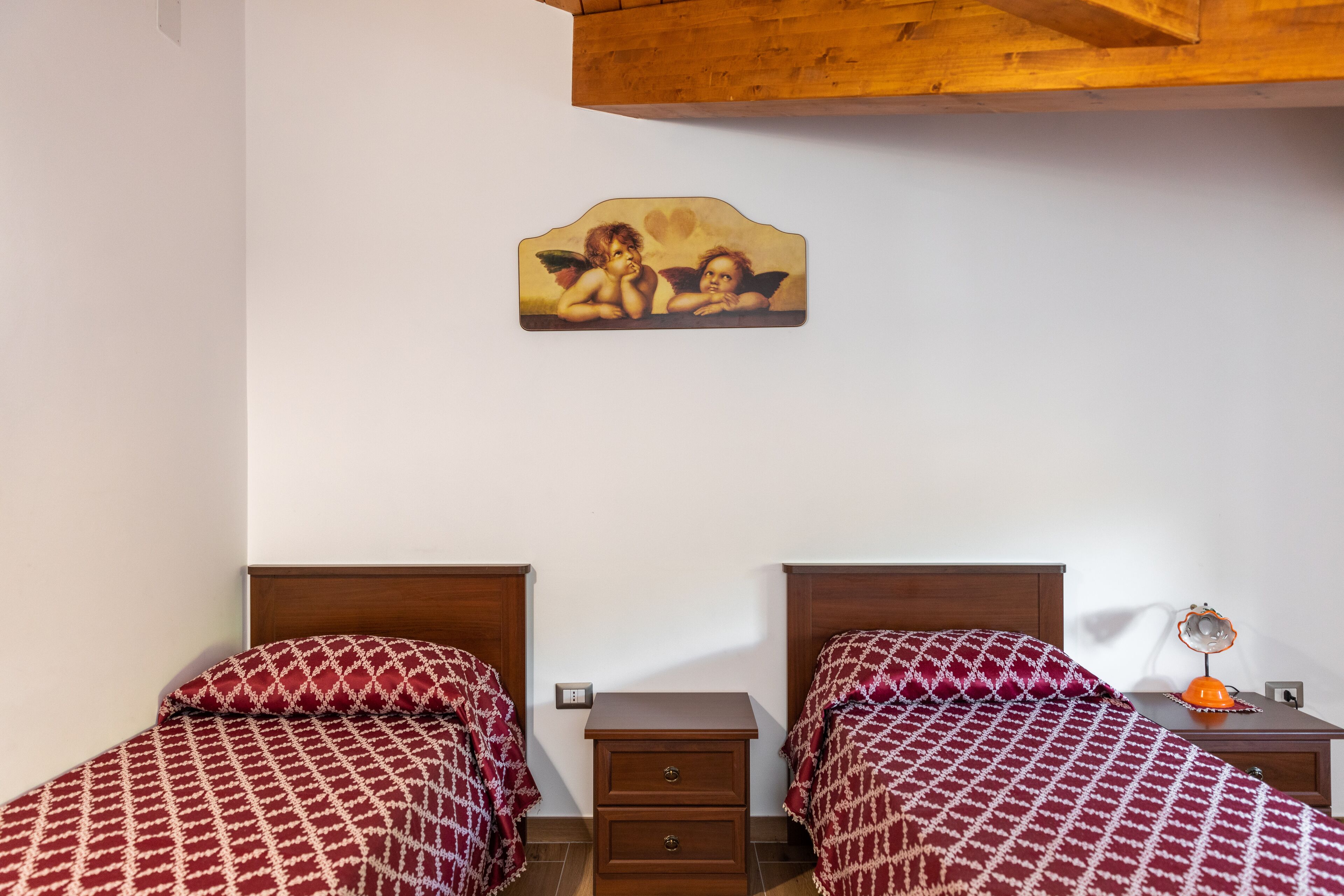 A bedroom with two beds, wooden headboards, a nightstand with a lamp, and a wall-mounted picture.