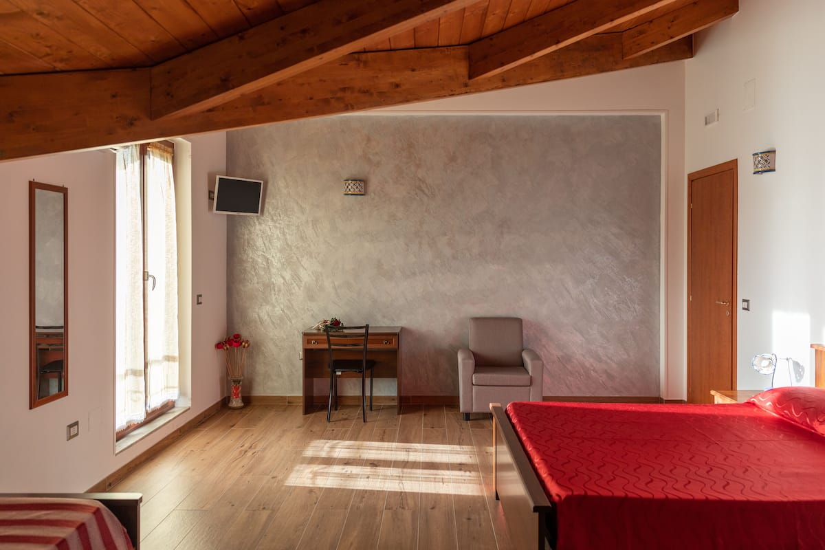 Ein Schlafzimmer mit Holzboden, einem Bett mit roter Decke, einem Schreibtisch mit Stuhl, einem an der Wand montierten Fernseher und einem Fenster mit Vorhängen.