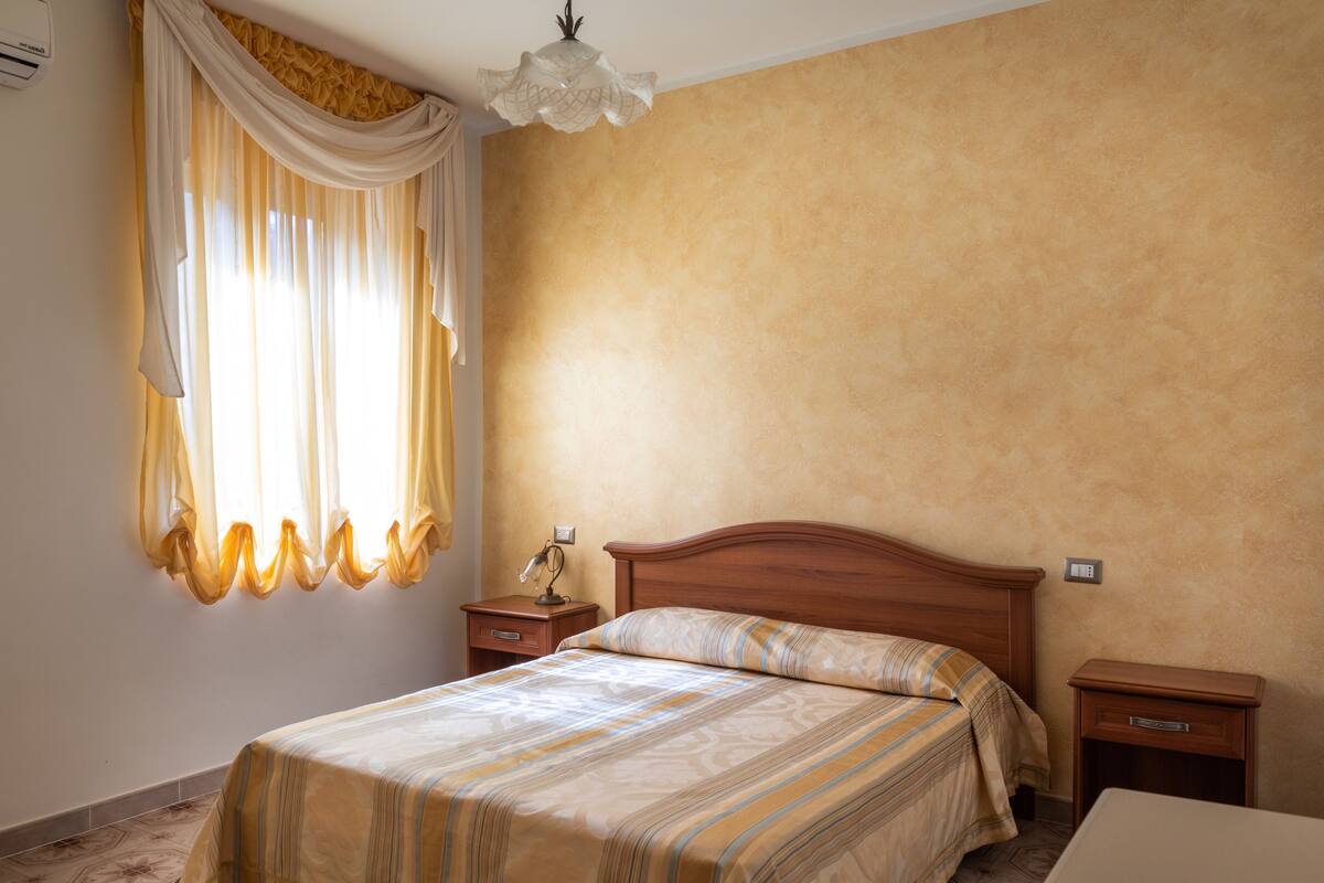 Ein Schlafzimmer mit einem Bett, Nachttischen, einer Lampe und einem Fenster mit Vorhängen.
