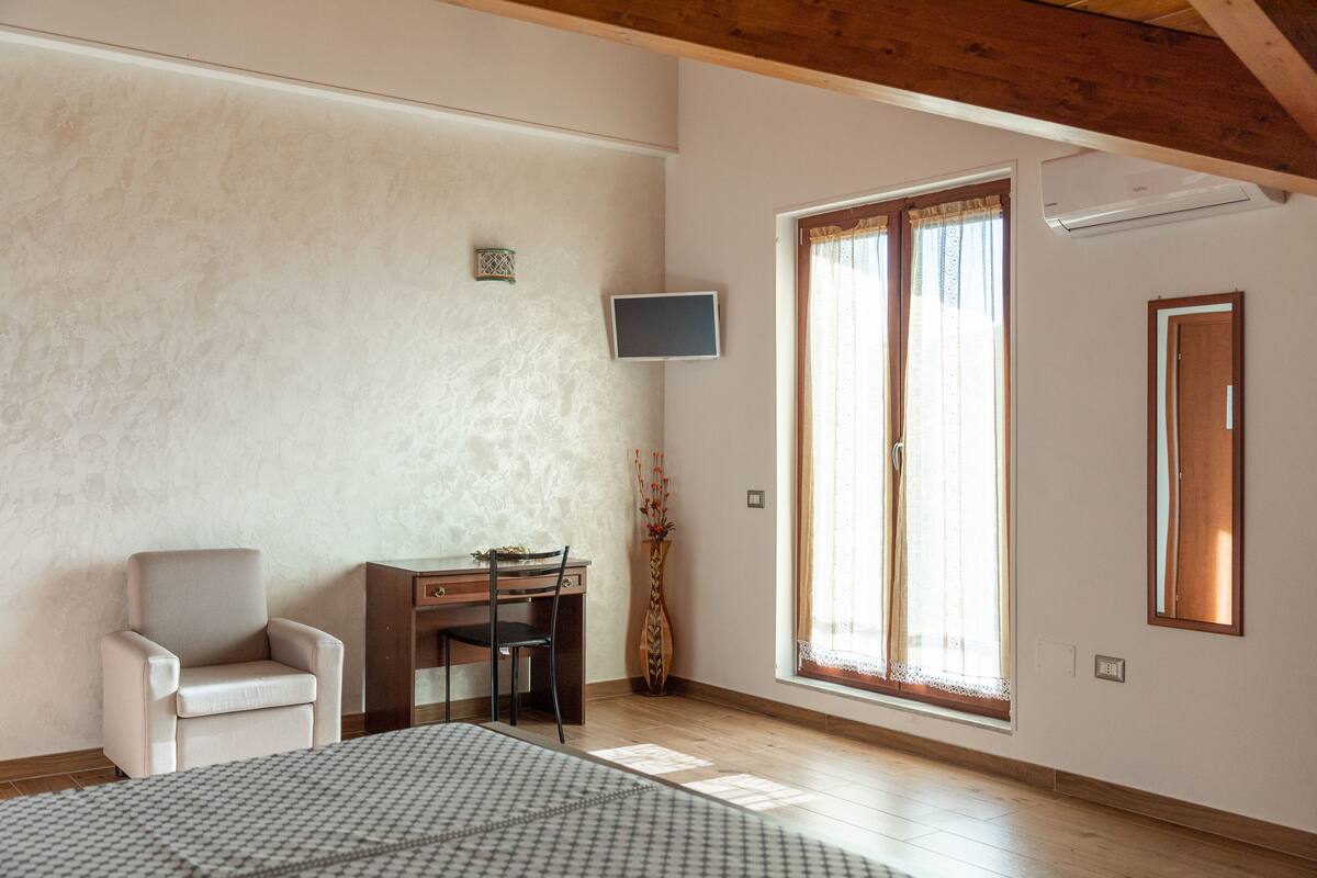 Ein Zimmer mit Holzboden, einem Schreibtisch mit Stuhl, einem an der Wand montierten Fernseher und einem großen Fenster mit Vorhängen.