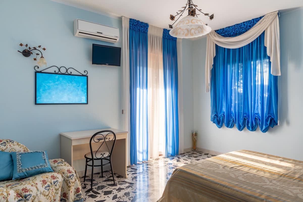 Ein Schlafzimmer mit einem Bett, einem Schreibtisch, einem Sessel, einem Fernseher und einem Fenster mit blauen Vorhängen.