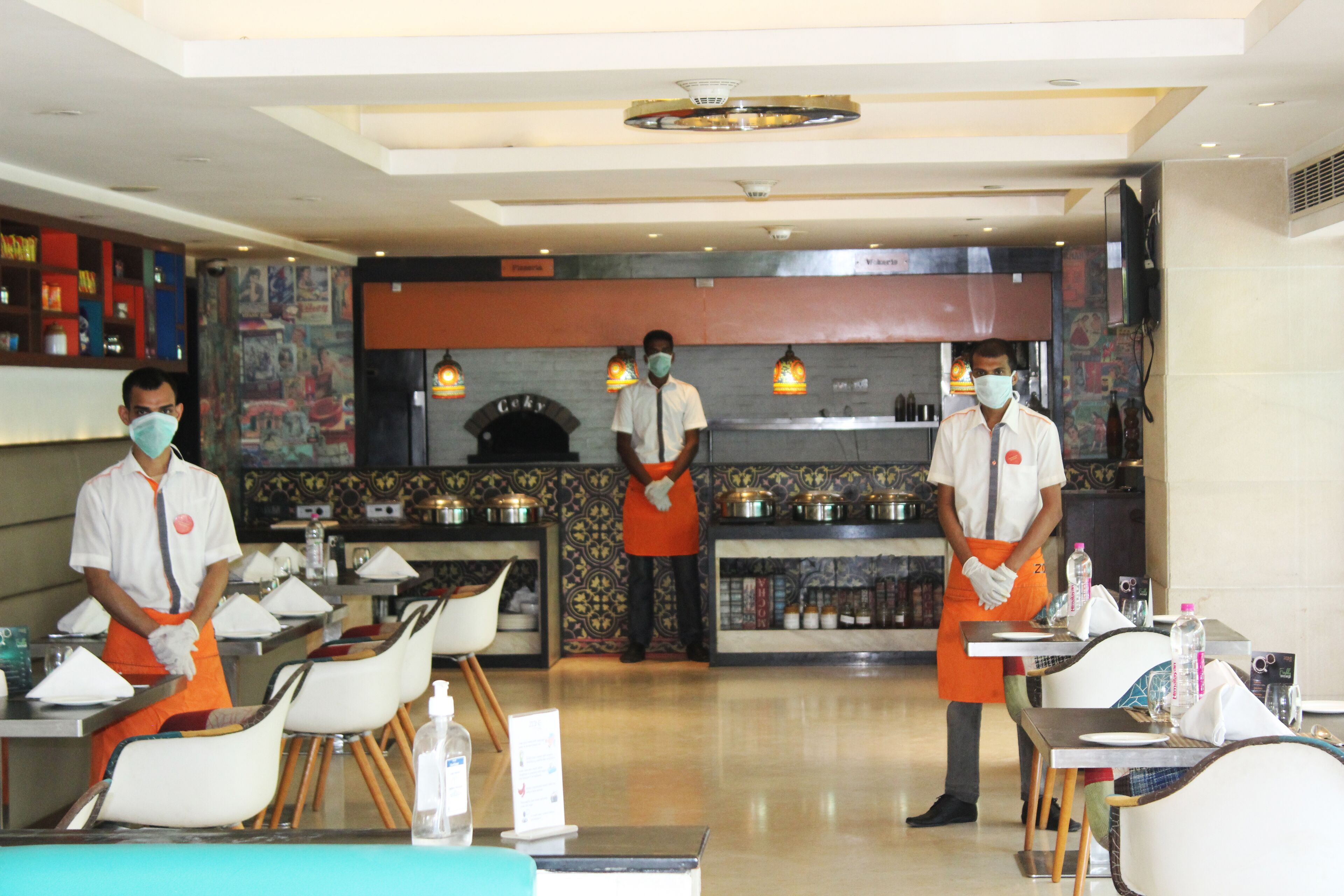 A restaurant with three waiters in uniform, tables set with white chairs, and a decorative wall with a fireplace.