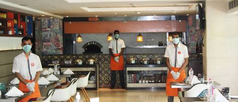 A restaurant with three waiters in uniform, tables set with white chairs, and a decorative wall with a fireplace.