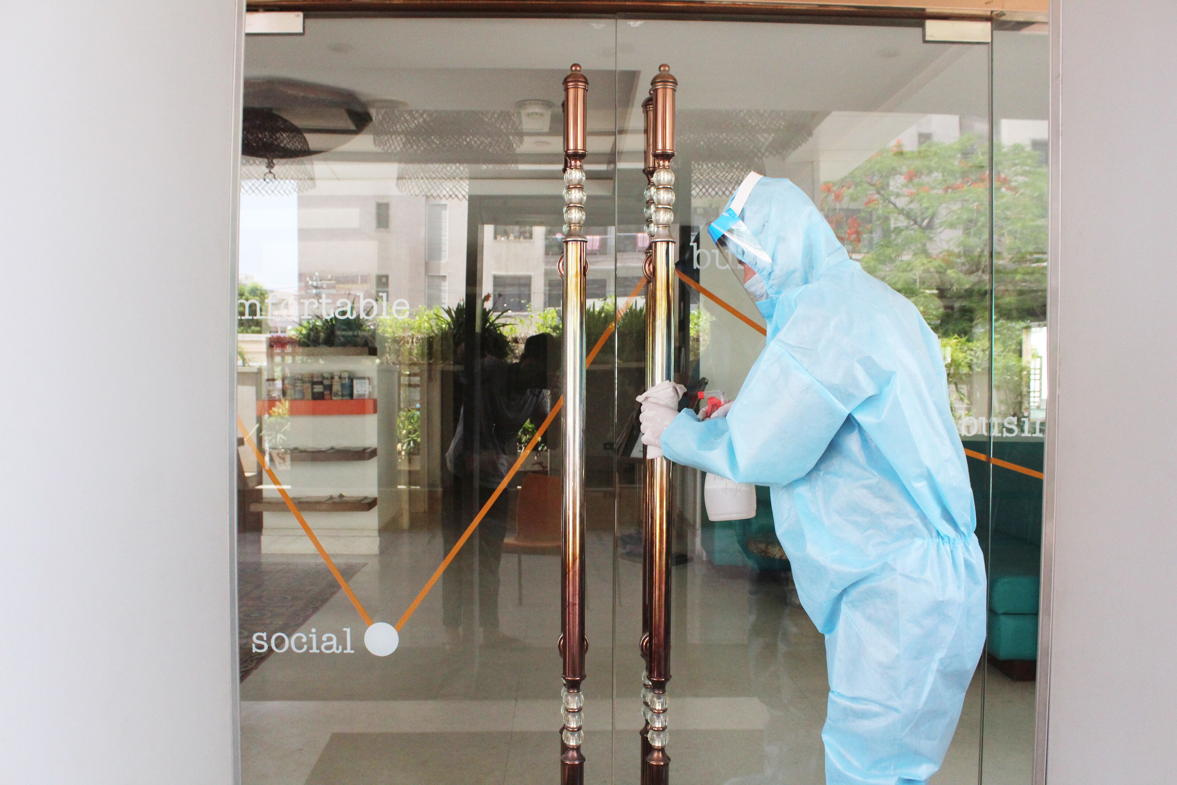 A person in a blue protective suit standing in front of a glass door with a reflection of a reception area and a plant.