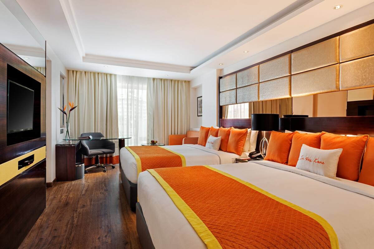 A hotel room with two beds, a TV, a desk, and a large window with curtains.