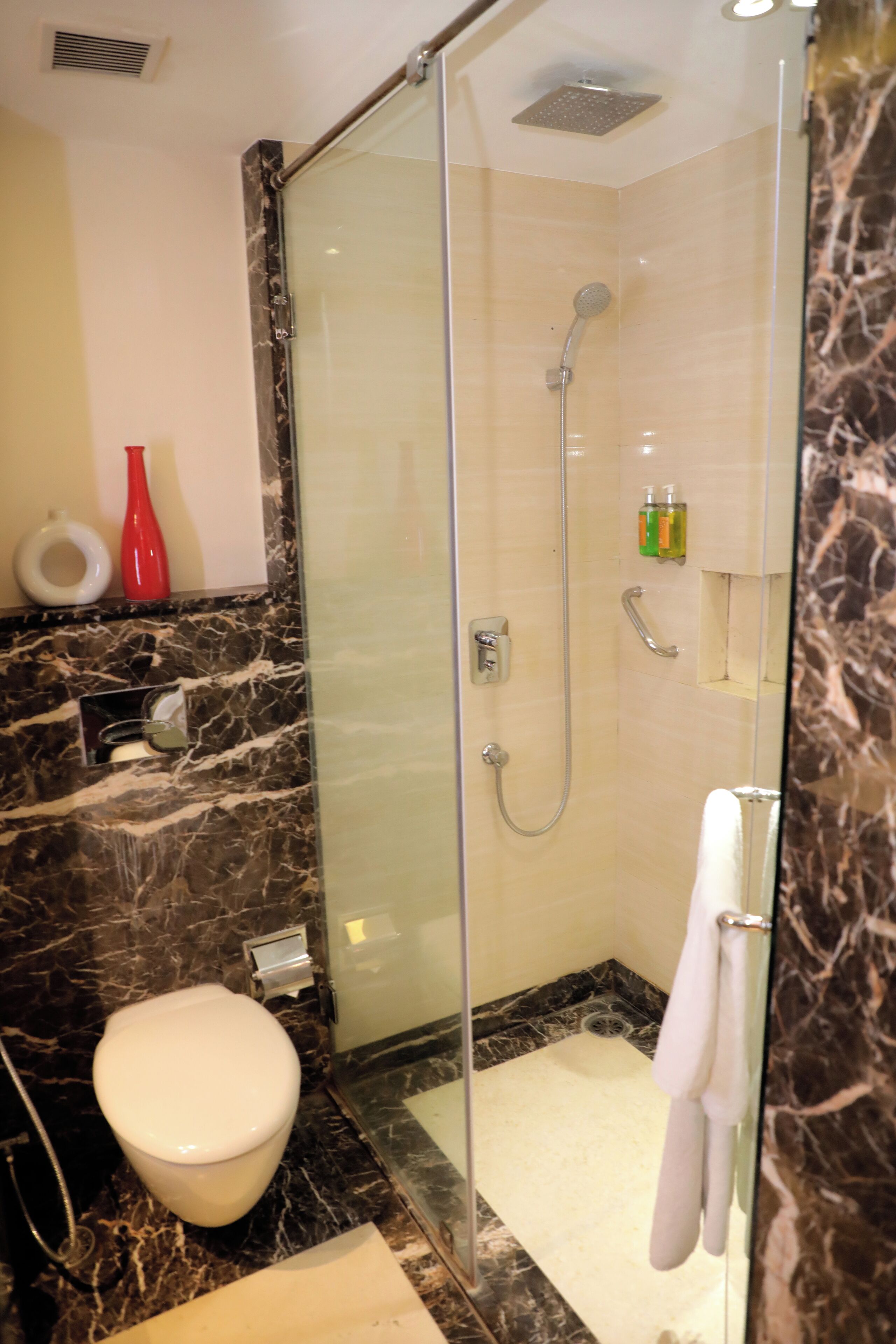 A modern bathroom with a glass-enclosed shower, a marble-tiled floor, a toilet, and a red vase on the counter.