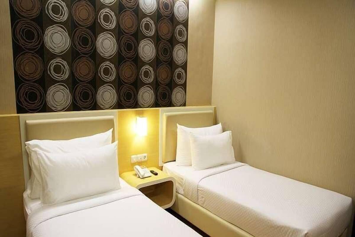 A hotel room with two single beds, a small bedside table with a lamp, and a patterned wallpaper above the beds.