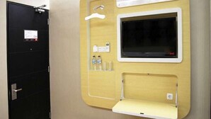 Habitació compacte amb una porta negra, un televisor penjat, una petita estant amb ampolles i una taula de nit blanca.