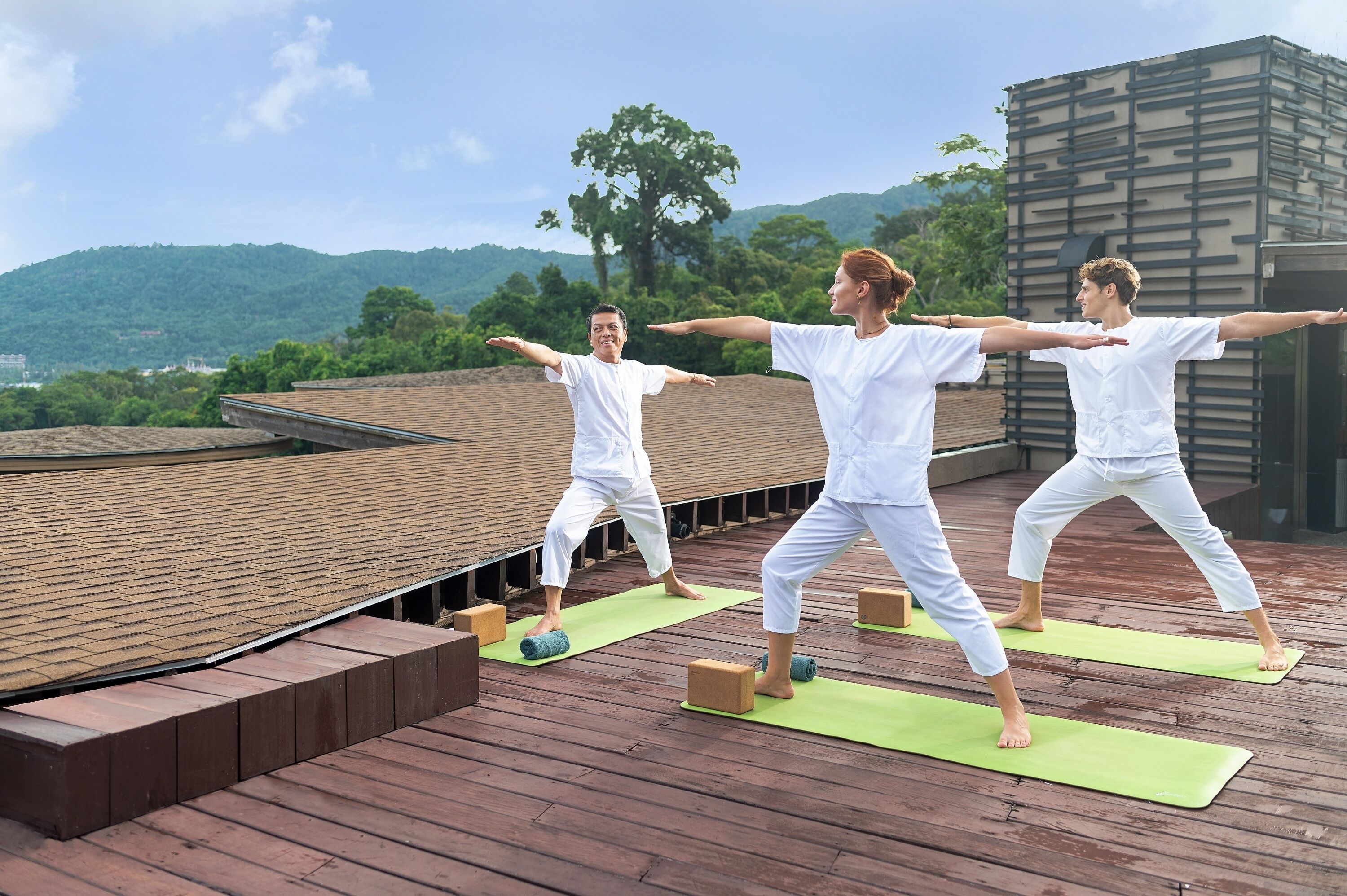 Tres personas practicando yoga en una terraza de madera con tapetes verdes, con vistas a un paisaje montañoso.