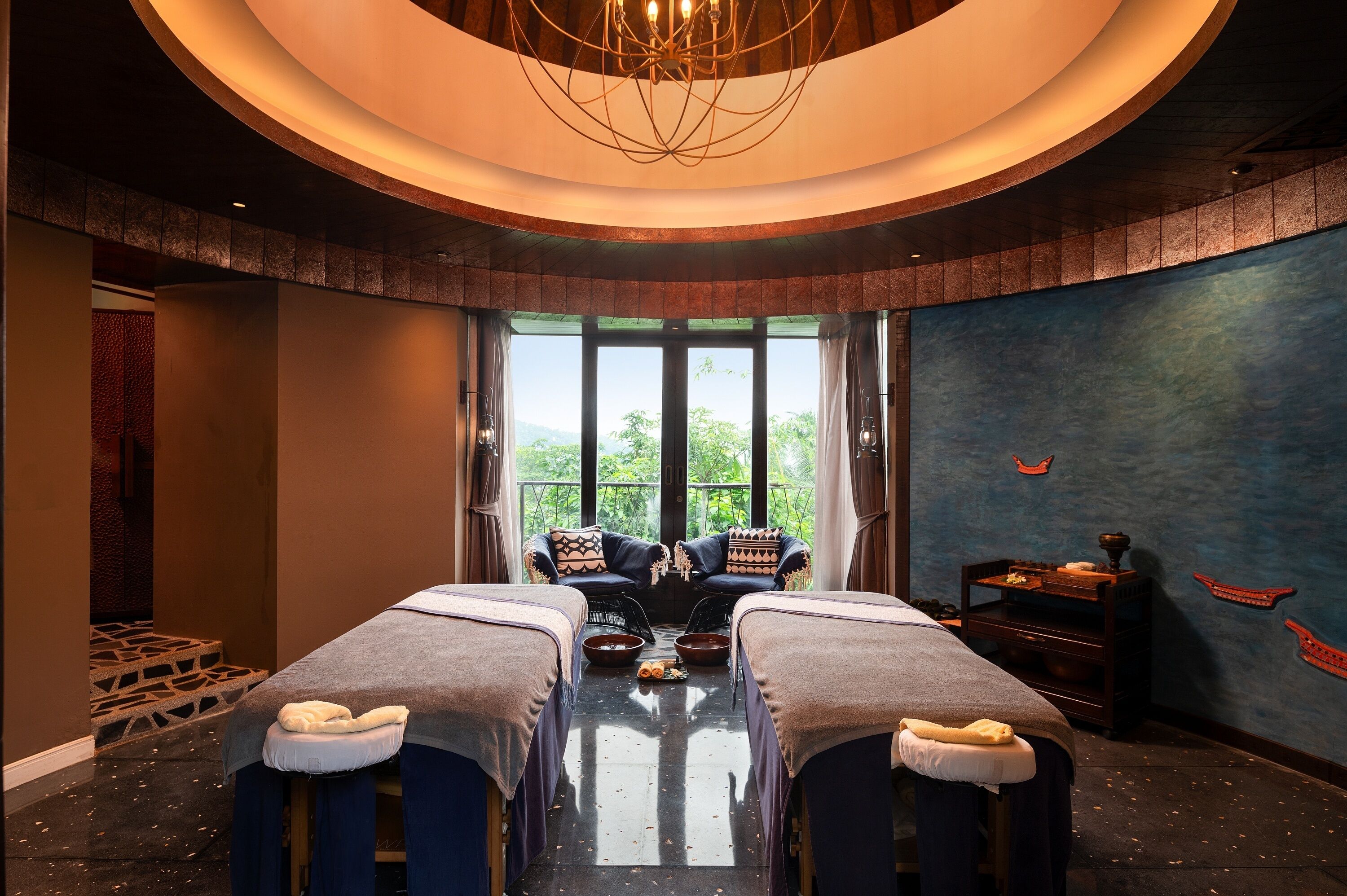 A room with two massage tables, a large window with a view, a ceiling light fixture, and a wall with a mural.