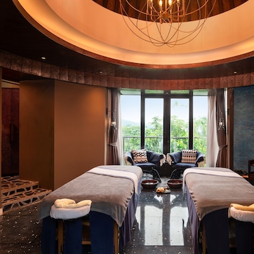 A room with two massage tables, a large window with a view, a ceiling light fixture, and a wall with a mural.