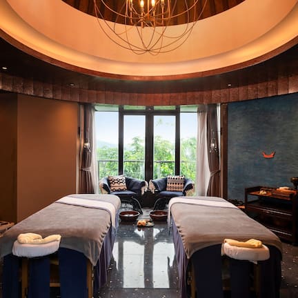 A room with two massage tables, a large window with a view, a ceiling light fixture, and a wall with a mural.