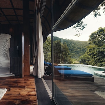 A room with a wooden floor, a glass wall, and a view of a river and forest.