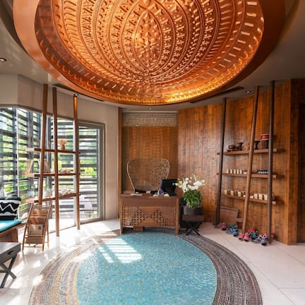 A room with a large circular ceiling design, a wooden desk, a seating area with cushions, and a view through large windows.