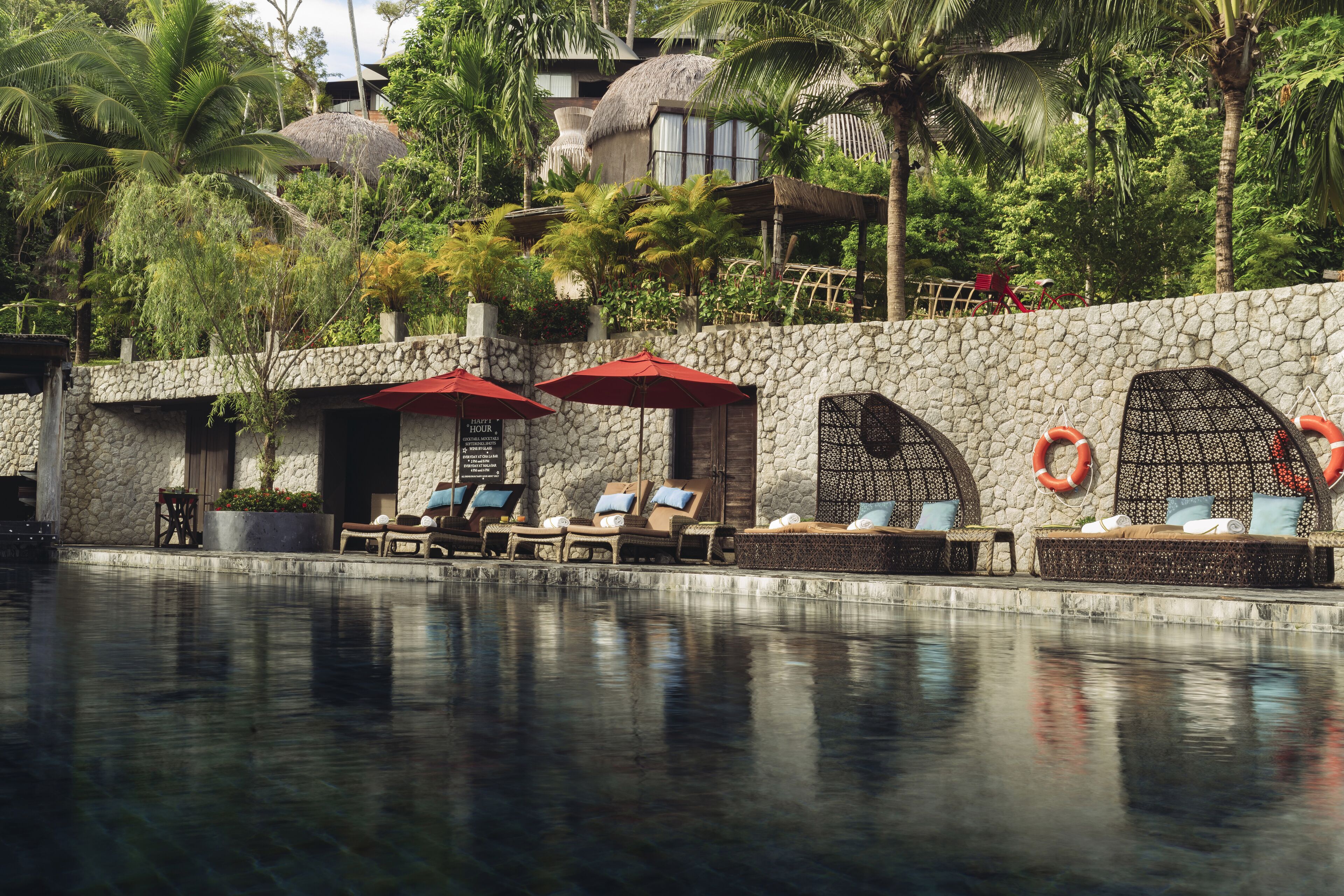 A poolside area with lounge chairs and umbrellas, surrounded by a stone wall and tropical vegetation.