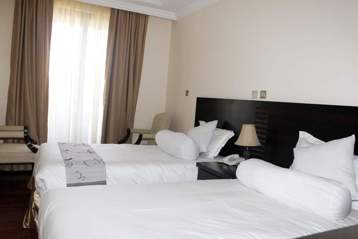 A hotel room with two beds, a chair, a bedside table with a lamp, and a window with curtains.