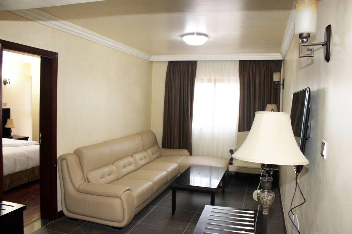 A hotel room with a beige sofa, a small table, a lamp, a TV, and a bed in the background.