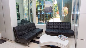 Uma área de espera com dois sofás de couro preto e uma mesa de centro branca, janelas amplas com vista para o exterior e uma parede de vidro refletindo o interior.
