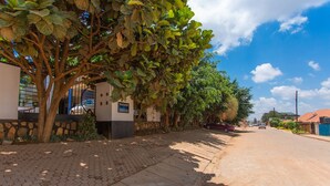 Uma rua pavimentada ladeada por árvores e edifícios, céu claro, e alguns carros estacionados.