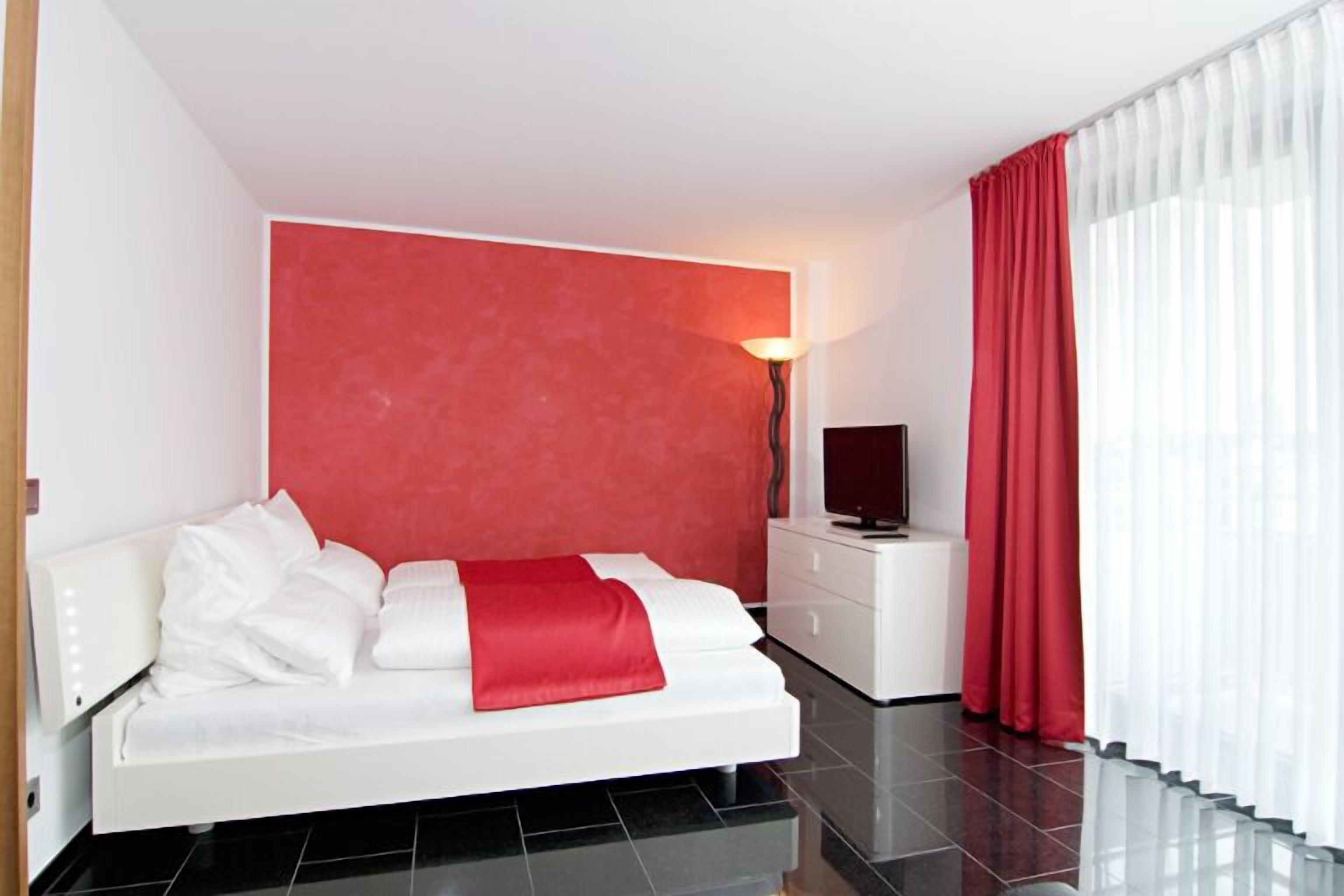 Ein modernes Hotelzimmer mit einem weißen Bett, einer roten Akzentwand, einem Flachbildfernseher und einem Glasgeländer am Fenster.