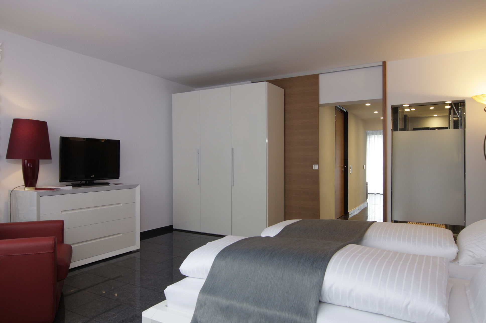 Ein modernes Hotelzimmer mit einem Bett, einem Fernseher auf einer weißen Kommode, einem roten Sessel und einem großen Spiegel.