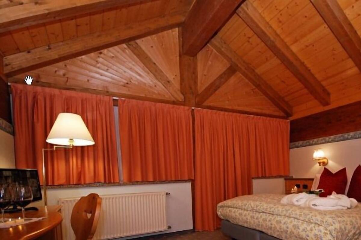 A hotel room with a wooden ceiling, a bed with red and white pillows, a bedside table with a lamp, and a radiator.