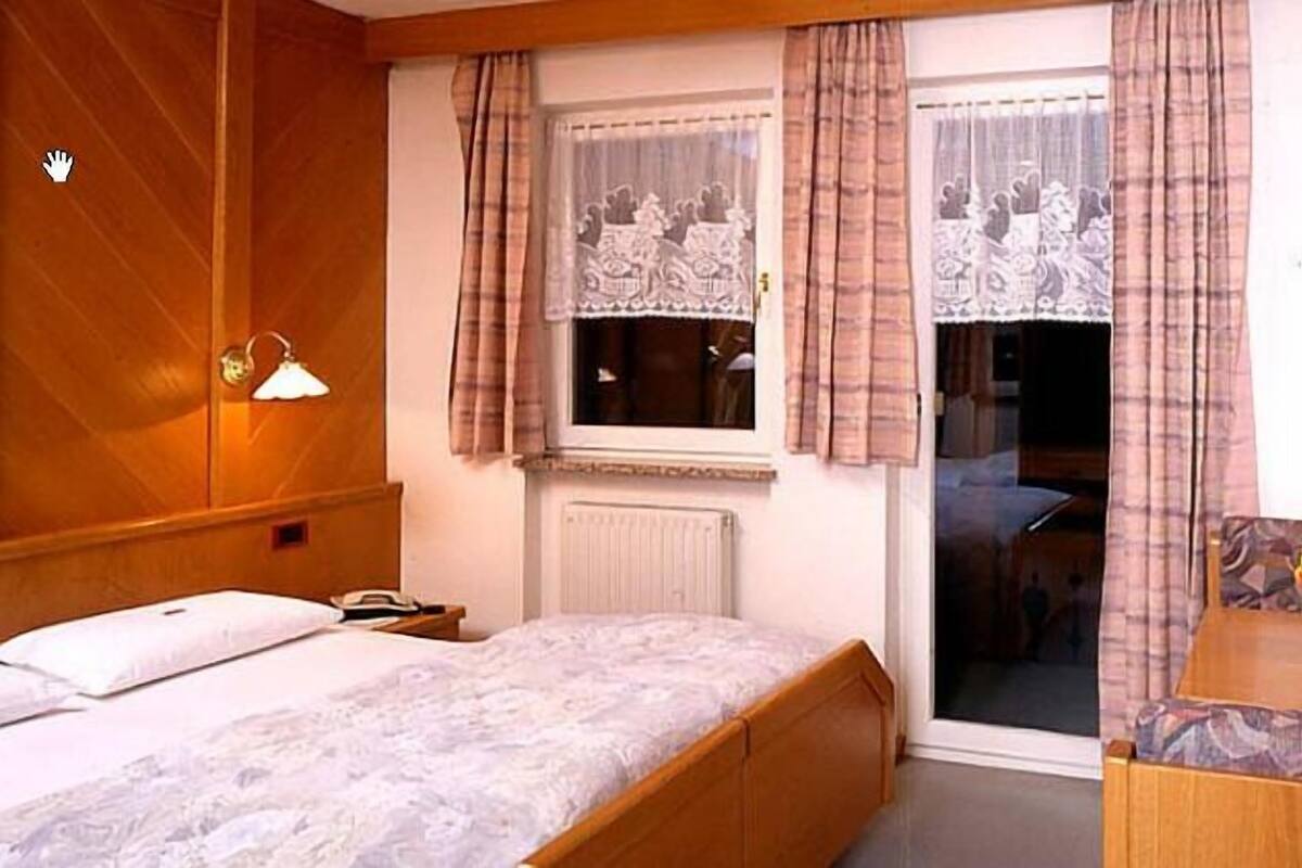 A bedroom with a bed, a bedside table with a lamp, a window with curtains, and a view of another room.