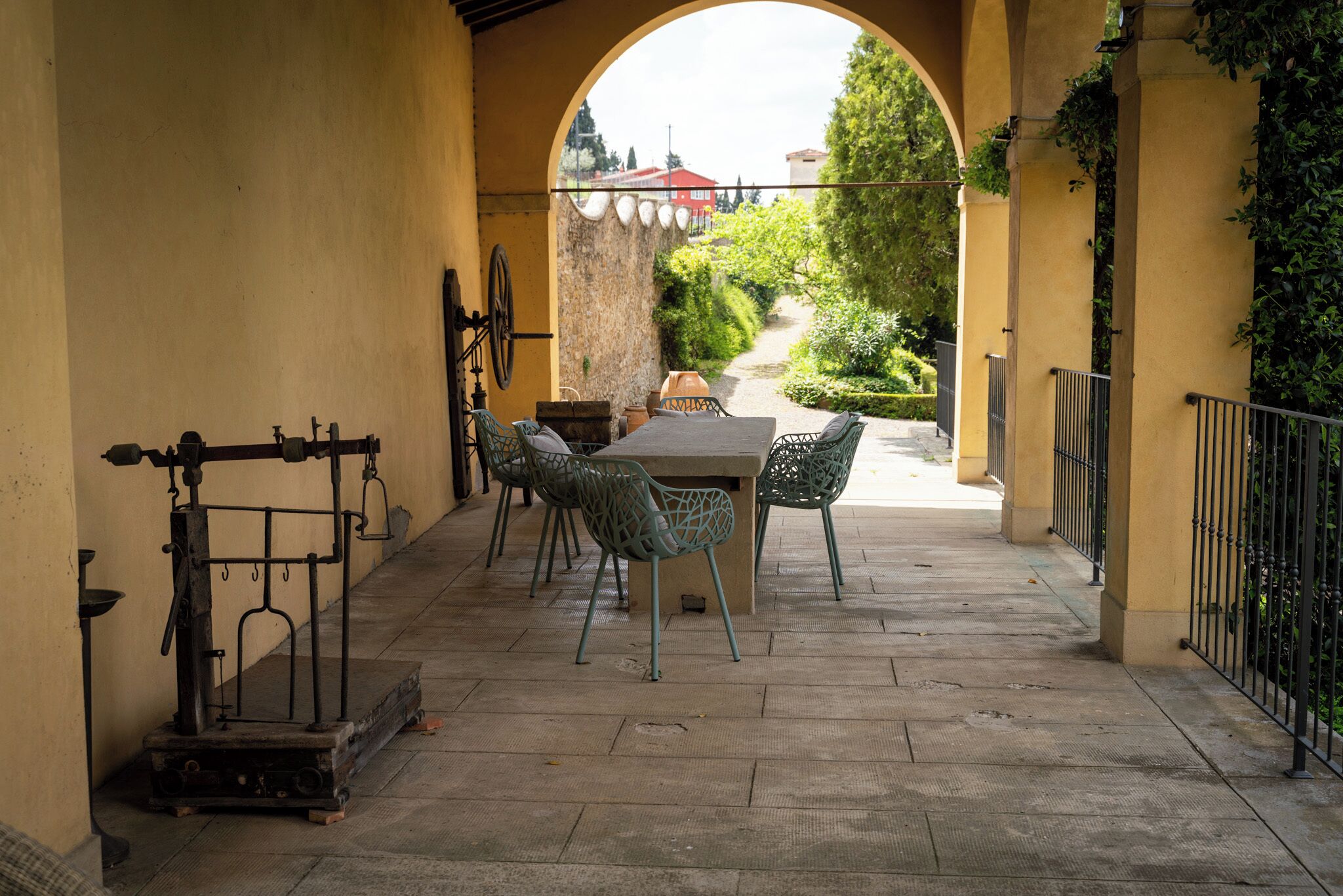 An outdoor patio with a table and chairs, a vintage water pump, and a view through an archway to a garden and buildings.
