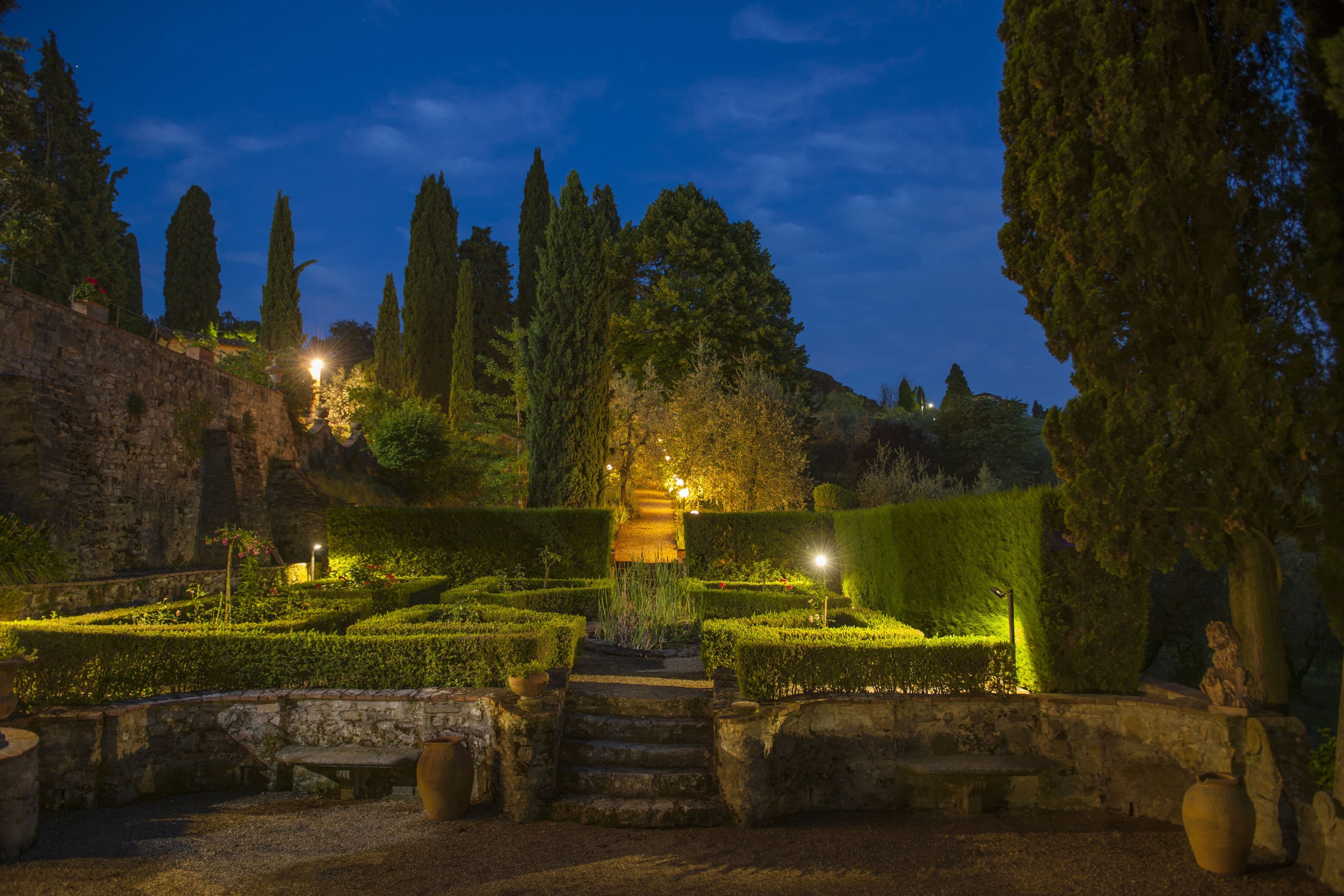 A well-maintained garden with a stone pathway leading to a central area, surrounded by neatly trimmed hedges and tall trees.
