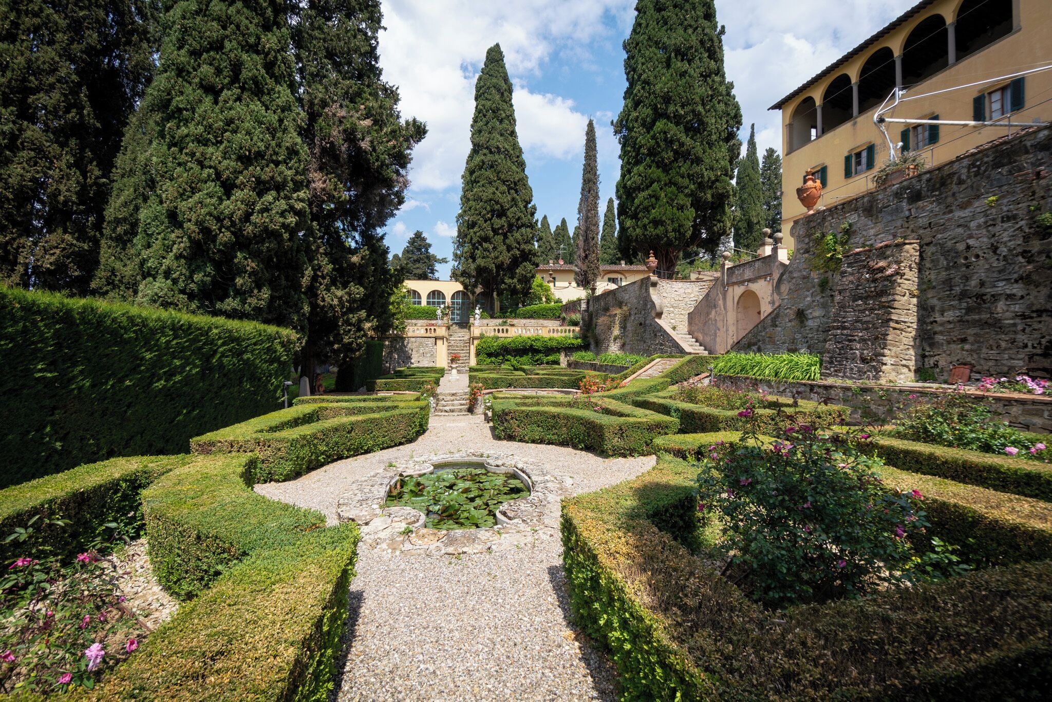 A formal garden with geometric hedges, a central pond, and tall cypress trees.