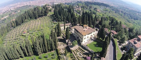 A large, historic building surrounded by lush gardens and tall trees, with a view of a distant cityscape.