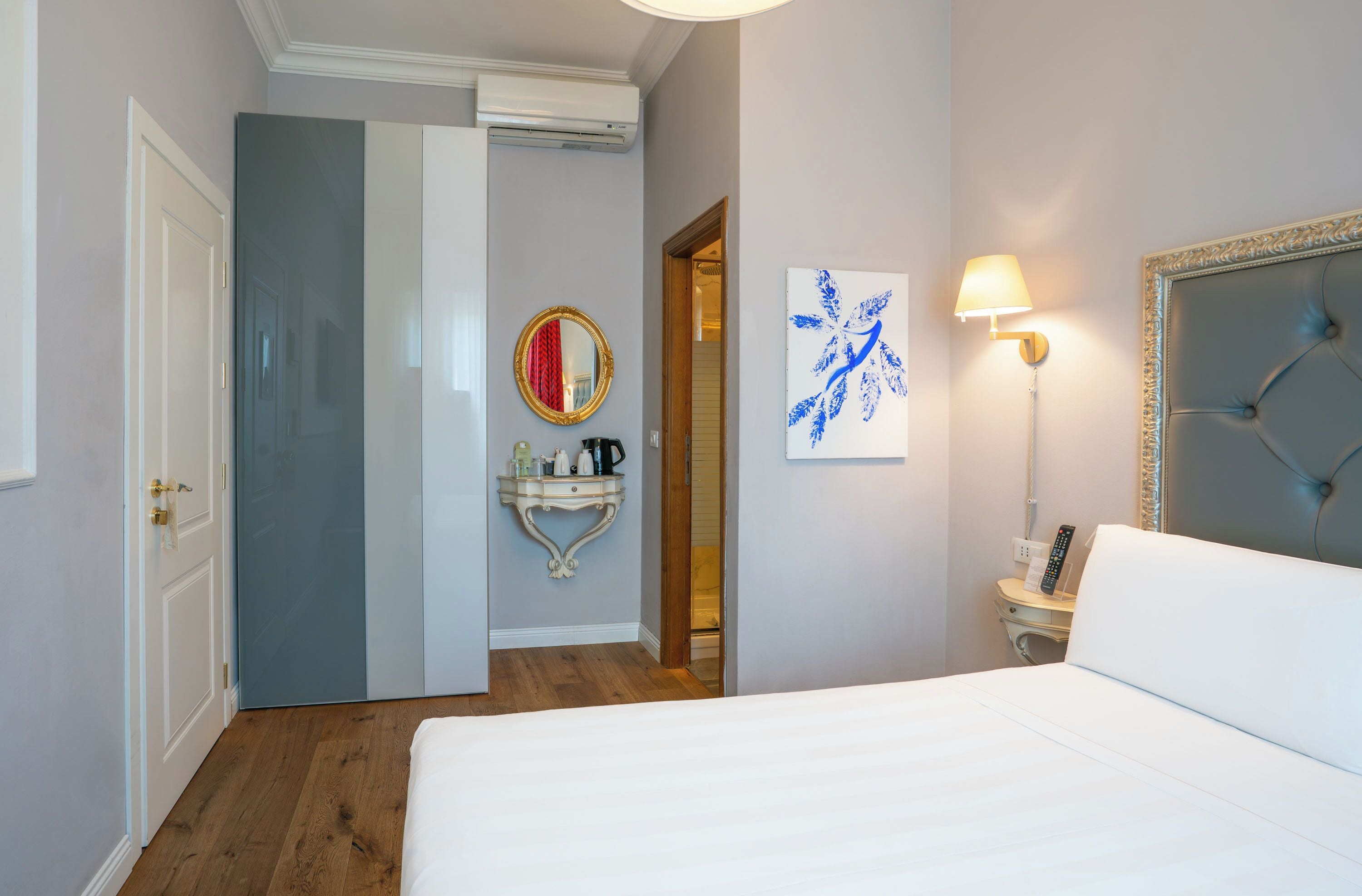 Ein Hotelzimmer mit einem Bett, einem Nachttisch mit Lampe, einem Wandspiegel, einem dekorativen Wandbild und einer Tür zu einem weiteren Raum.