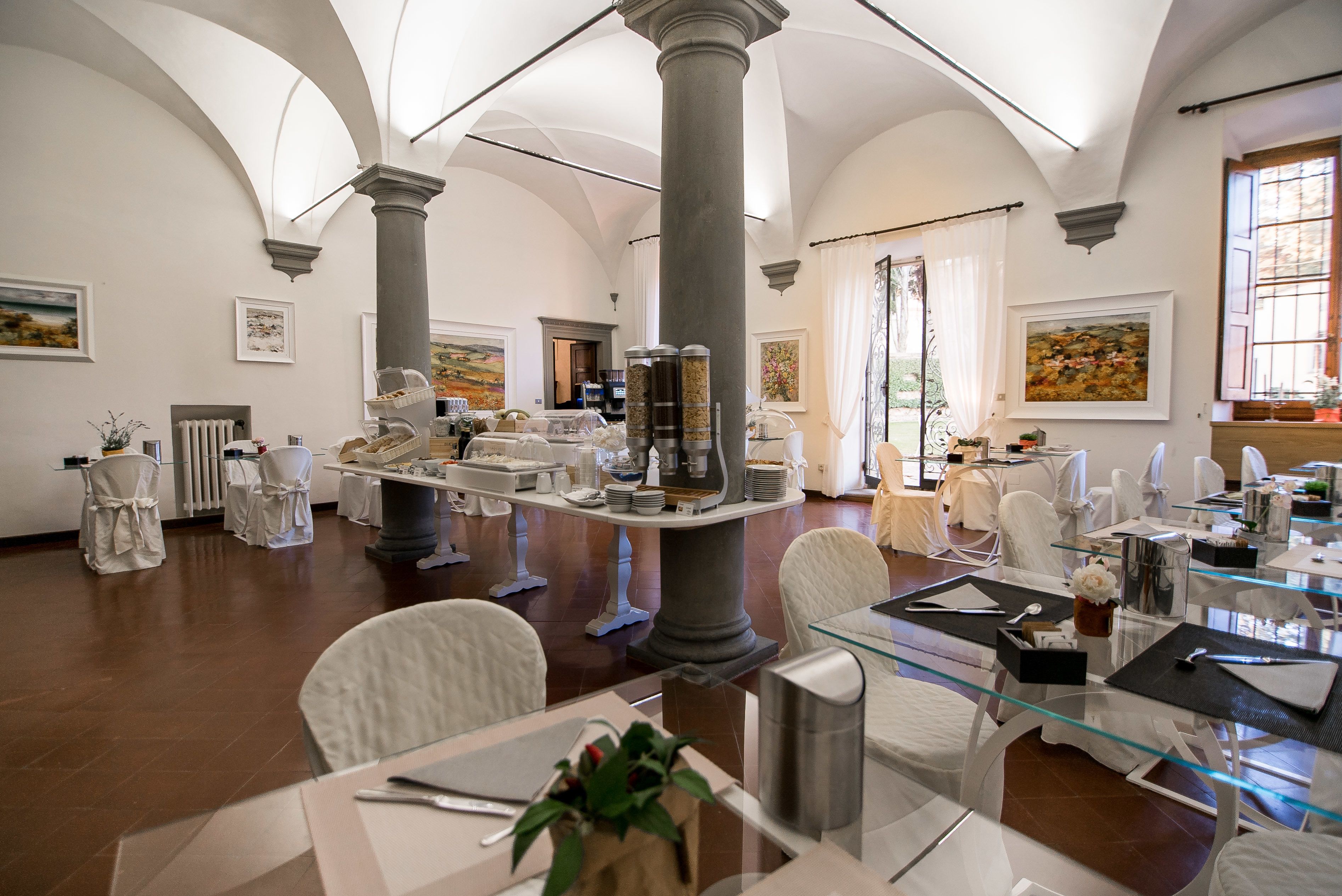 A spacious dining area with high ceilings, white walls, and large windows. Tables are set with white tablecloths and chairs, and there are several decorative items and paintings on the walls.