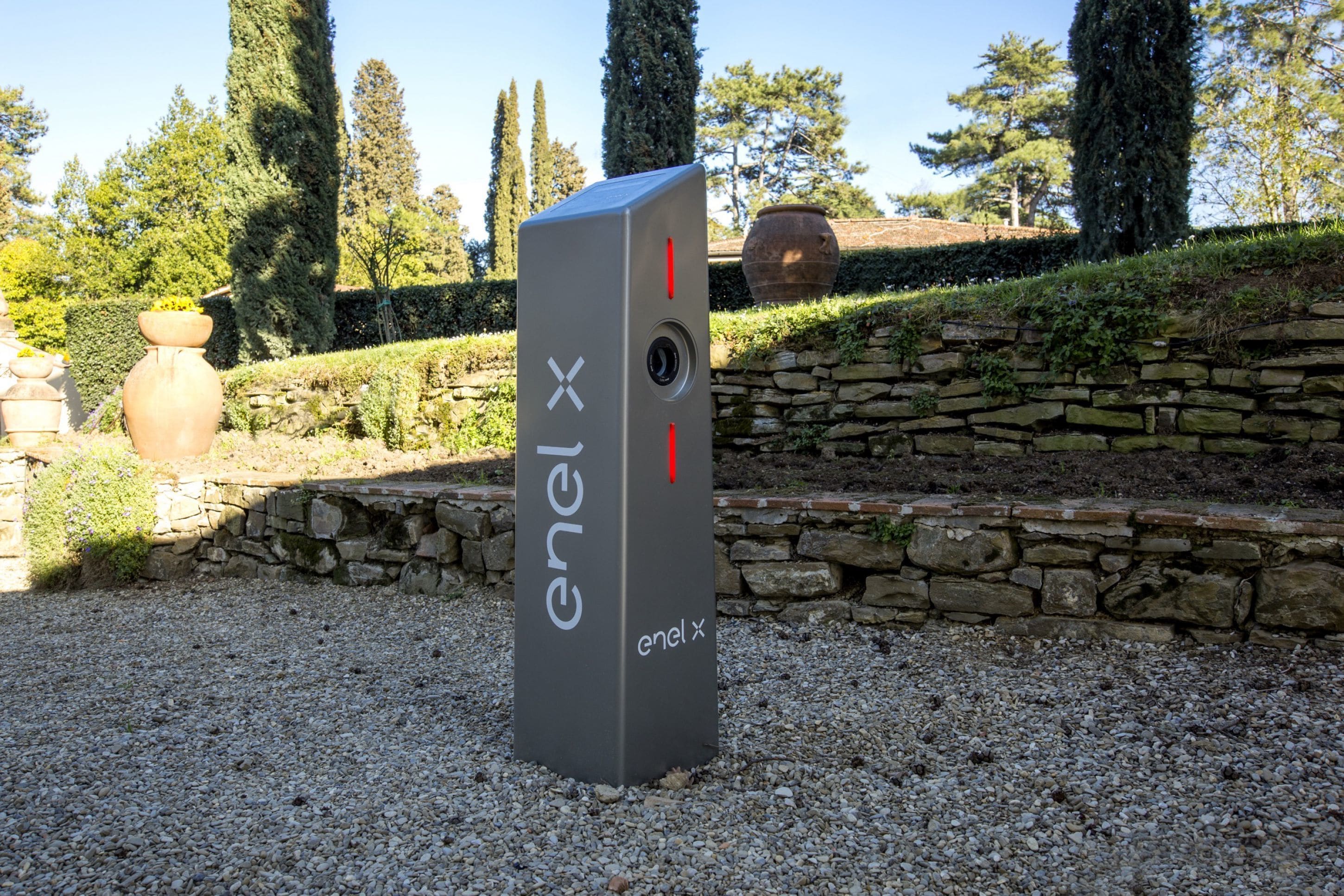 A grey ENEL X energy storage unit in a garden setting with stone walls and trees.