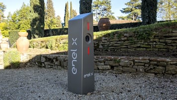 A grey ENEL X energy storage unit in a garden setting with stone walls and trees.